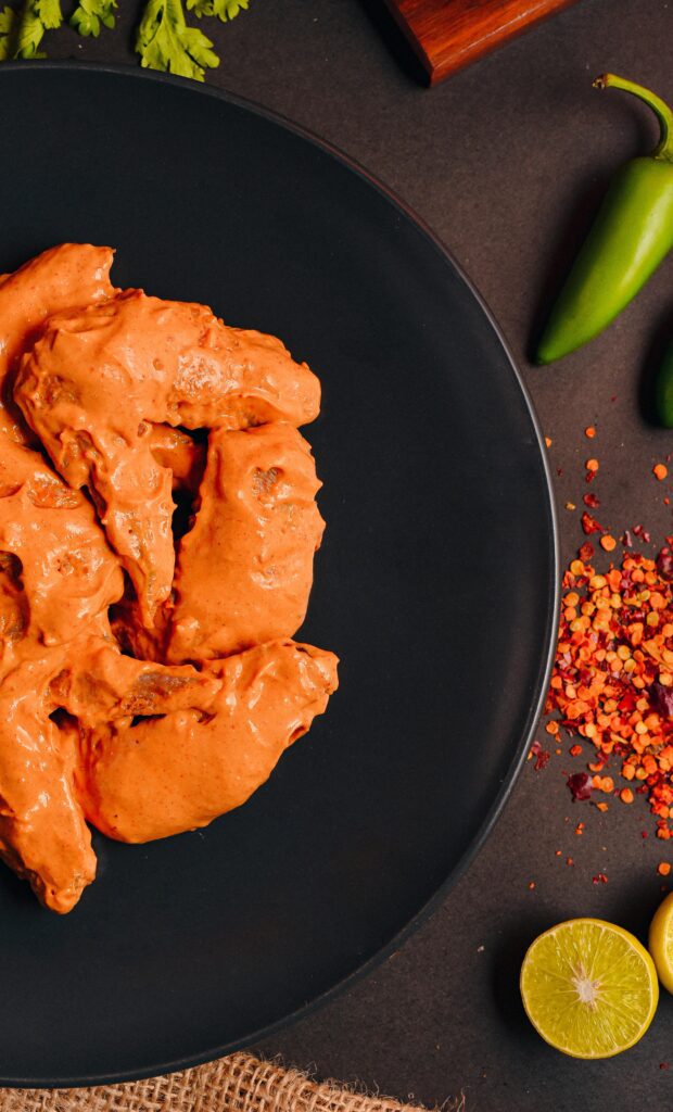 Marinated tandoori chicken wings on a black plate with fresh lemon, chili flakes, and green chili.

