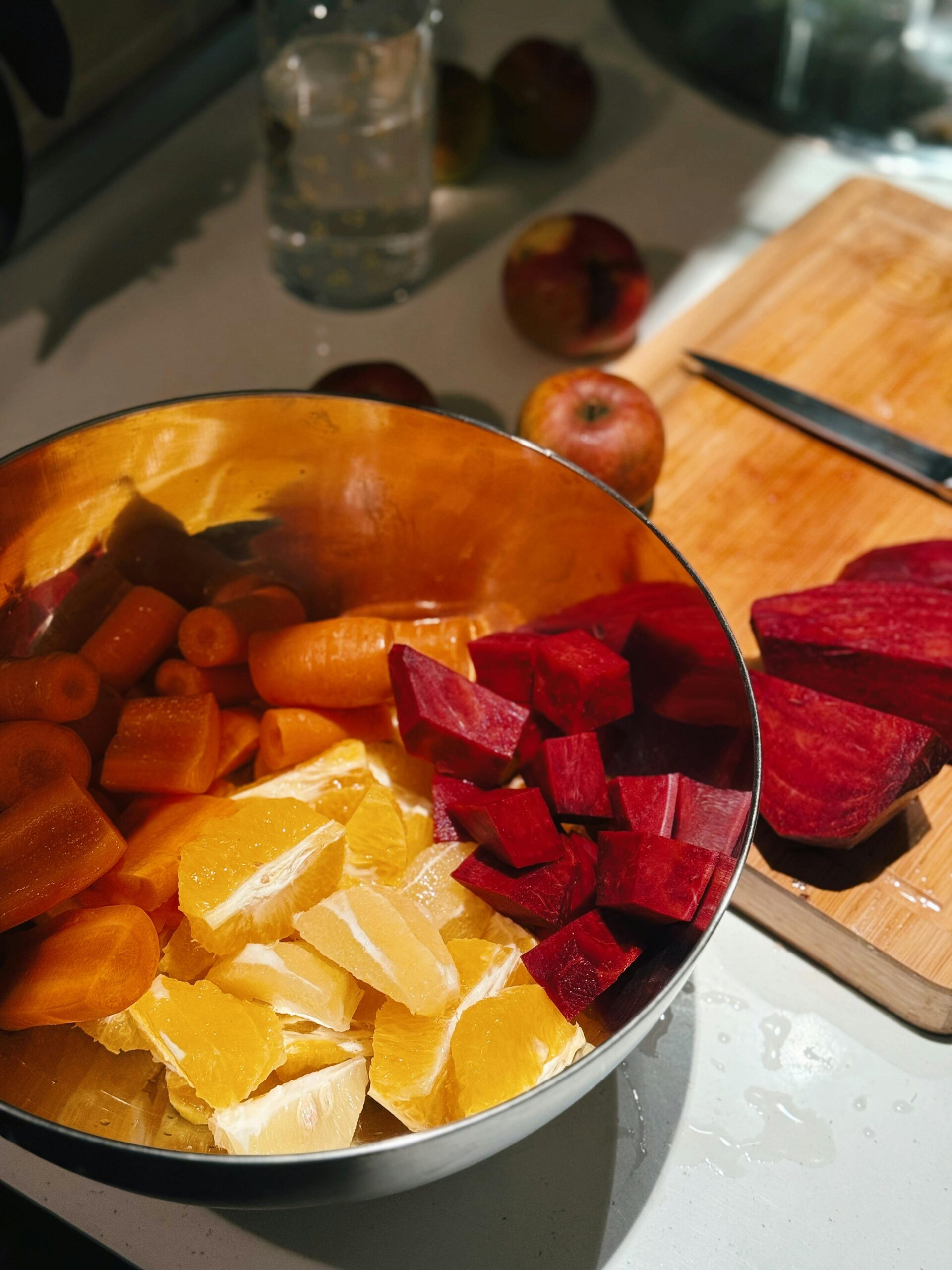 A metal bowl filled with chopped carrots, beetroots, and orange slices on a kitchen counter with a wooden cutting board.