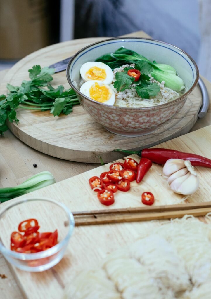 A bowl of homemade chicken ramen with fresh bok choy, soft-boiled eggs, and chili peppers on a wooden cutting board.