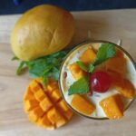 A refreshing glass of healthy mango lassi made with almond milk, topped with fresh mango chunks, a cherry, and mint leaves.