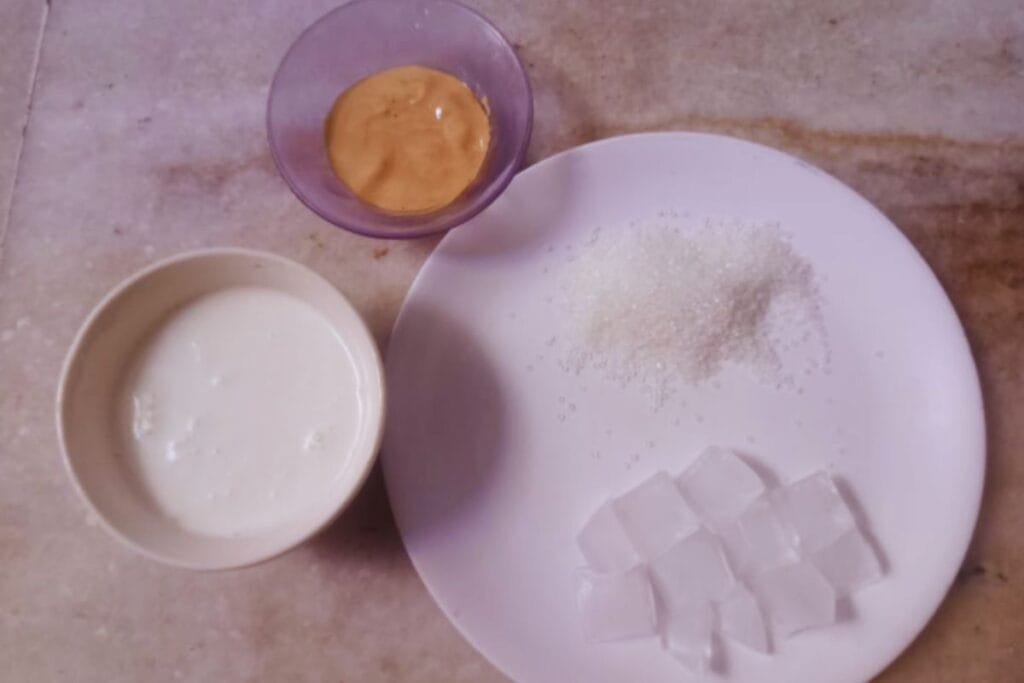 Top-down view of coffee ingredients: a bowl of milk, a small bowl of coffee powder, and ice cubes on a white plate.